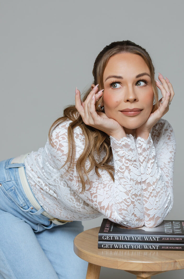 Julie Solomon in a white lace top leaning on a stool beside her book
